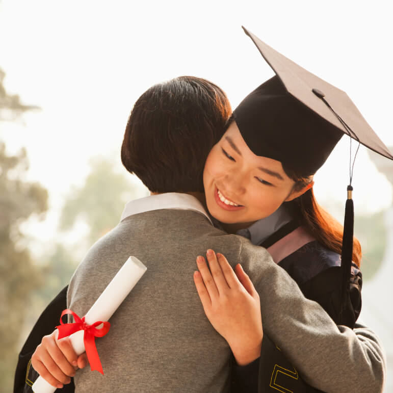 A student wearing a graduation hat and holding a diploma hugging their parent